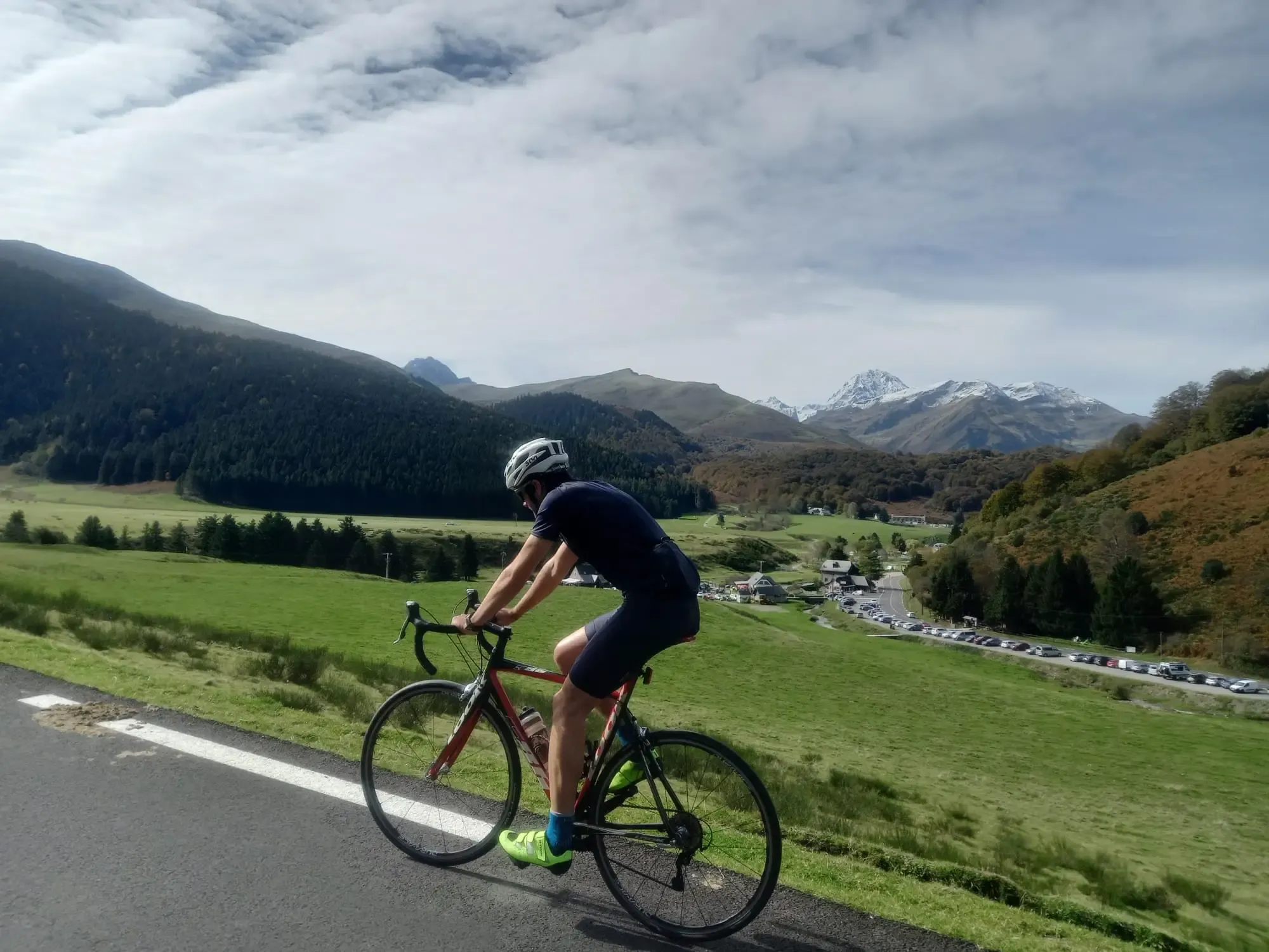 Cycliste de route en pleine ascension de la face Ouest du Col d'Aspin à vélo. Le coureur, portant un maillot bleu, des chaussures vert fluo et un casque blanc, pédale sur la route de montagne sinueuse traversant les prairies verdoyantes des Pyrénées, avec des sommets enneigés et le massif du Pic du Midi en arrière-plan.